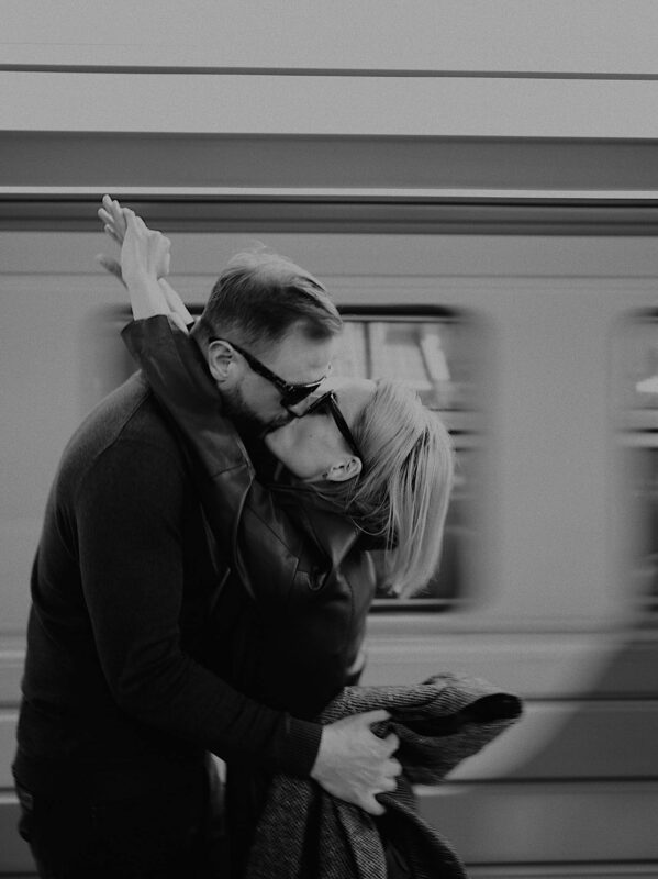 A couple shares a passionate kiss at a train station in black and white.